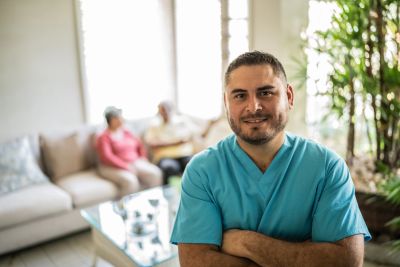 Smiling healthcare worker in scrubs with patients in the background.