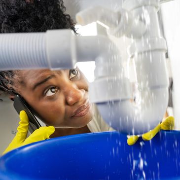 Woman in yellow gloves inspecting leaking pipe while talking on phone.