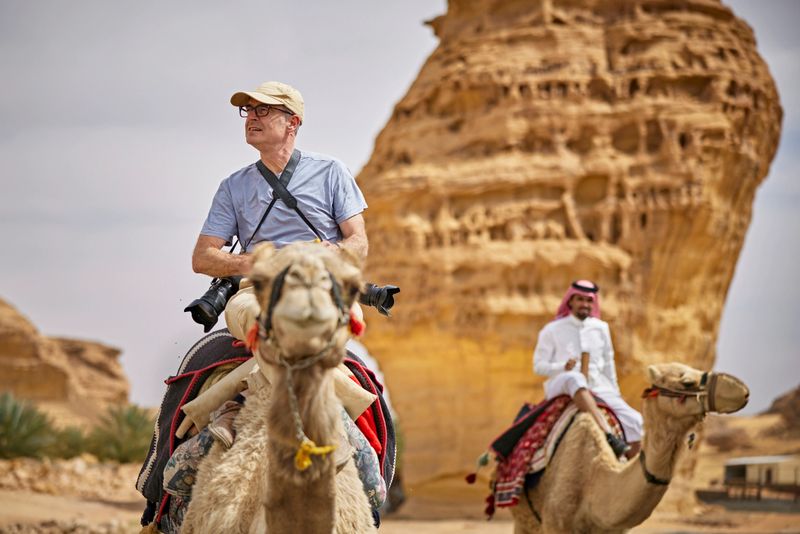 Focus on foreground man in early 50s exploring Al-Ula desert environment and sandstone formations with young Saudi man in traditional attire.