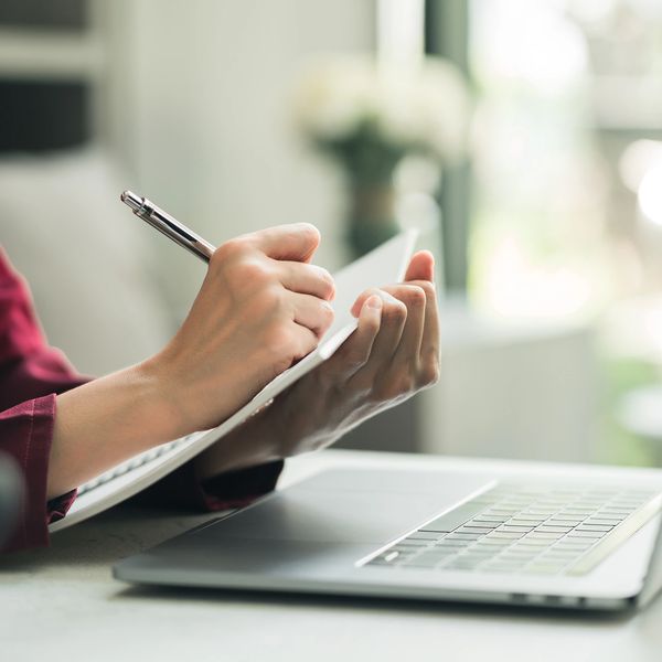 Person writing on a notepad beside an open laptop in a bright room.
