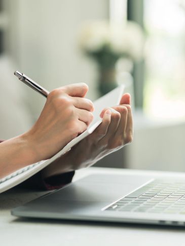 Person writing on a notepad beside an open laptop in a bright room.