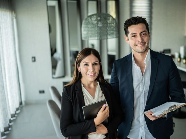 Two young professionals smiling indoors with tablet and clipboard.