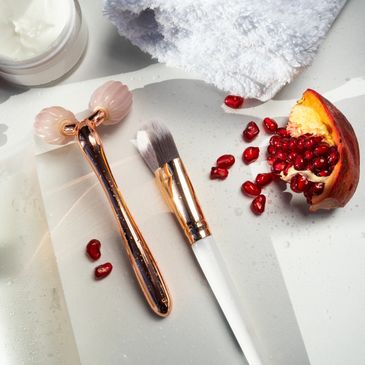 Beauty tools and pomegranate seeds arranged on a white surface with a towel.