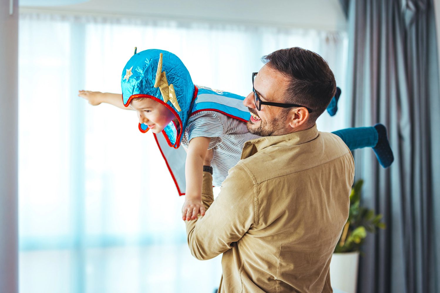 Father playfully lifts son dressed as a superhero, pretending to fly indoors.