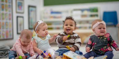 Four babies playing with colorful toys indoors.