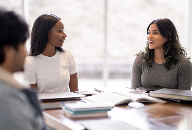 A diverse group of university students discuss their course material while sitting together around the conference table.