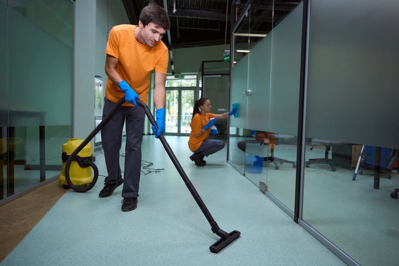Young man vacuuming the floor while his collegue cleaning the glass surface in the building