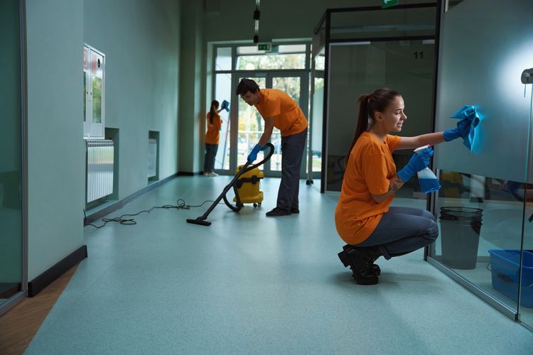 Three cleaners are cleaning in a office building. The cleaners are vacuuming and window cleaning.