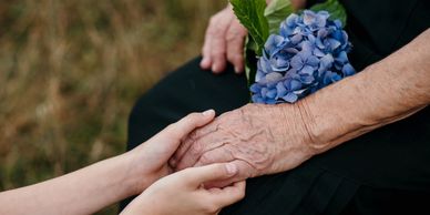Young hands gently holding an elderly hand with blue flowers on lap.