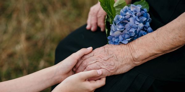 Young hands gently holding an elderly hand with blue flowers on lap.