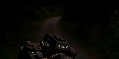 Hand holding a Sony camera on a dark, forest path at sunset.