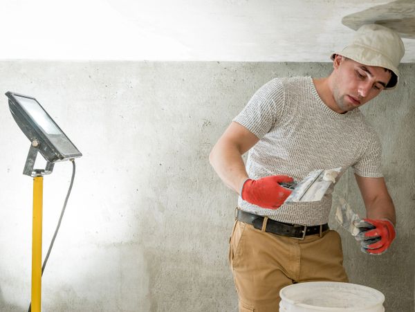Worker preparing plaster with tools near a bucket in a dimly lit room.