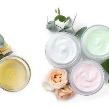 Jars of colorful skincare creams with flowers and leaves on white.