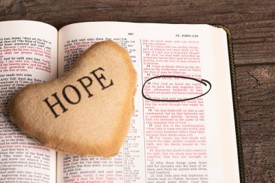 A heart-shaped stone with 'HOPE' rests on an open Bible highlighting a verse about hope and faith.