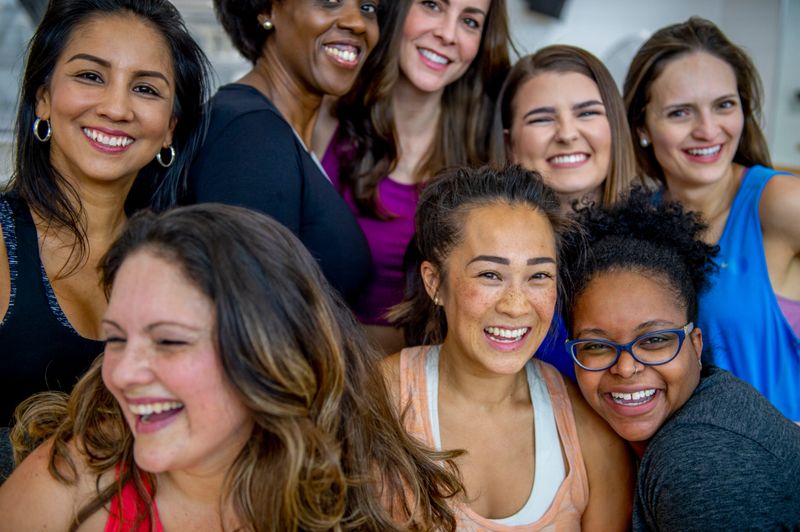 A large group of racially diverse middle aged woman, huddle together for a group portrait after a fitness class.  They are each dressed comfortably in athletic wear and are smiling as they laugh with one another.