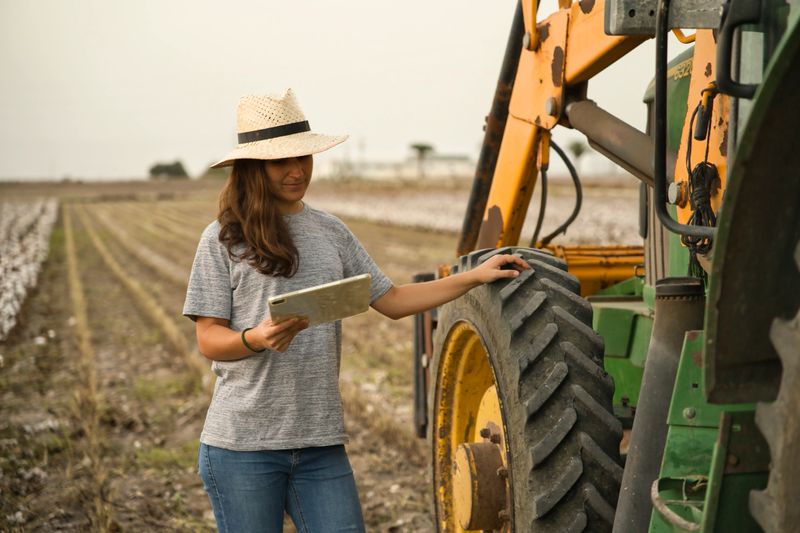 Portrait of smiling young woman farmer using tablet and tractor for harvest. Modern agriculture with the concept of technology and machinery