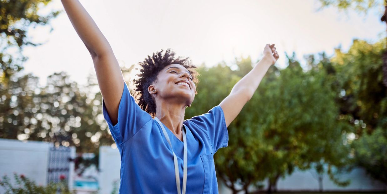 A joyful healthcare worker stretches arms outdoors in morning light.