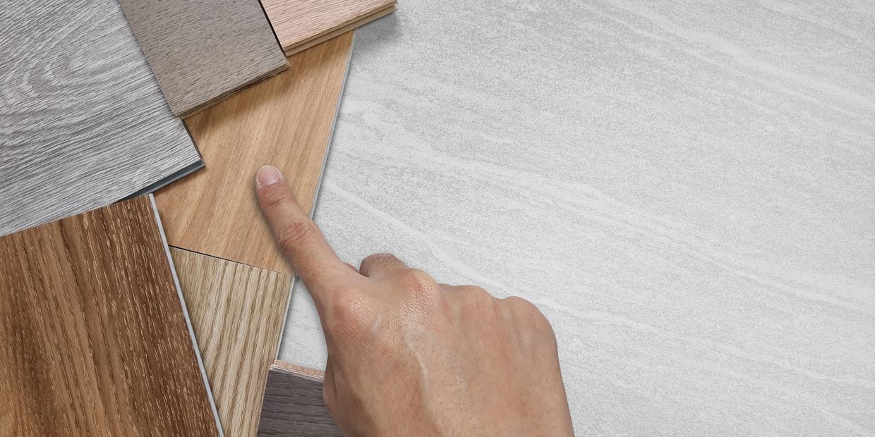 A hand pointing at various wood-look flooring samples on a light gray stone-look surface.