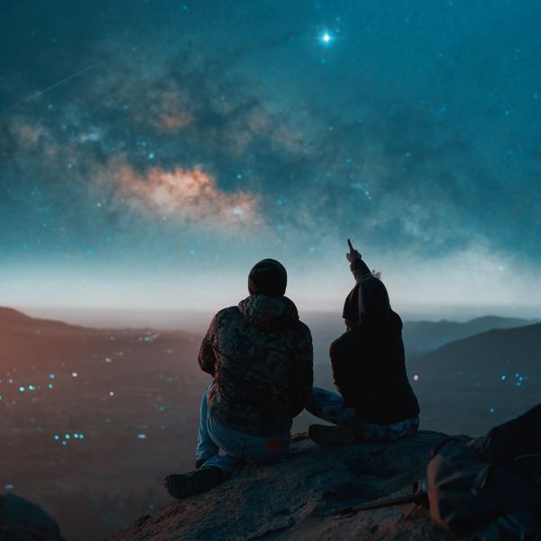 Two people stargazing on a mountain under a vibrant night sky.