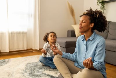 A mother and daughter meditate together in a cozy living room.