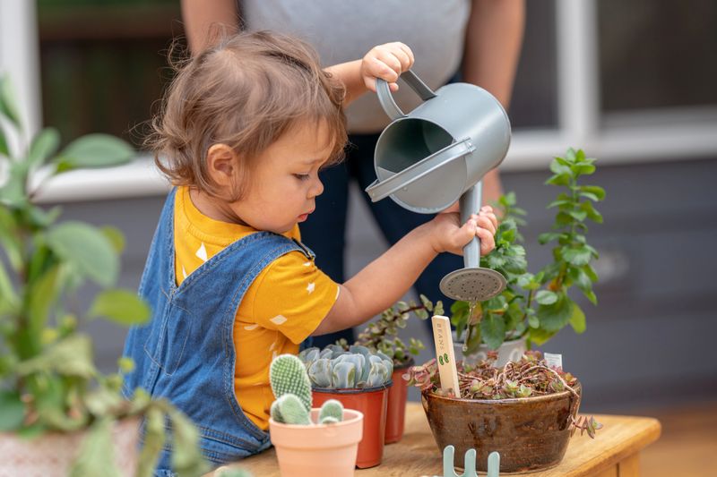 Cropped shot of a multiracial toddler girl using a watering can to water potted plants outside on the patio. The child is helping her mother with their garden.