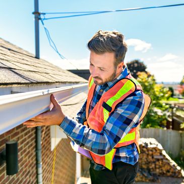 A man in a safety vest inspects a house gutter on a sunny day.