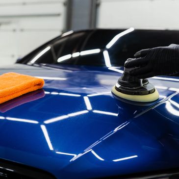 Hand polishing a blue car hood with an electric buffer and orange cloth.