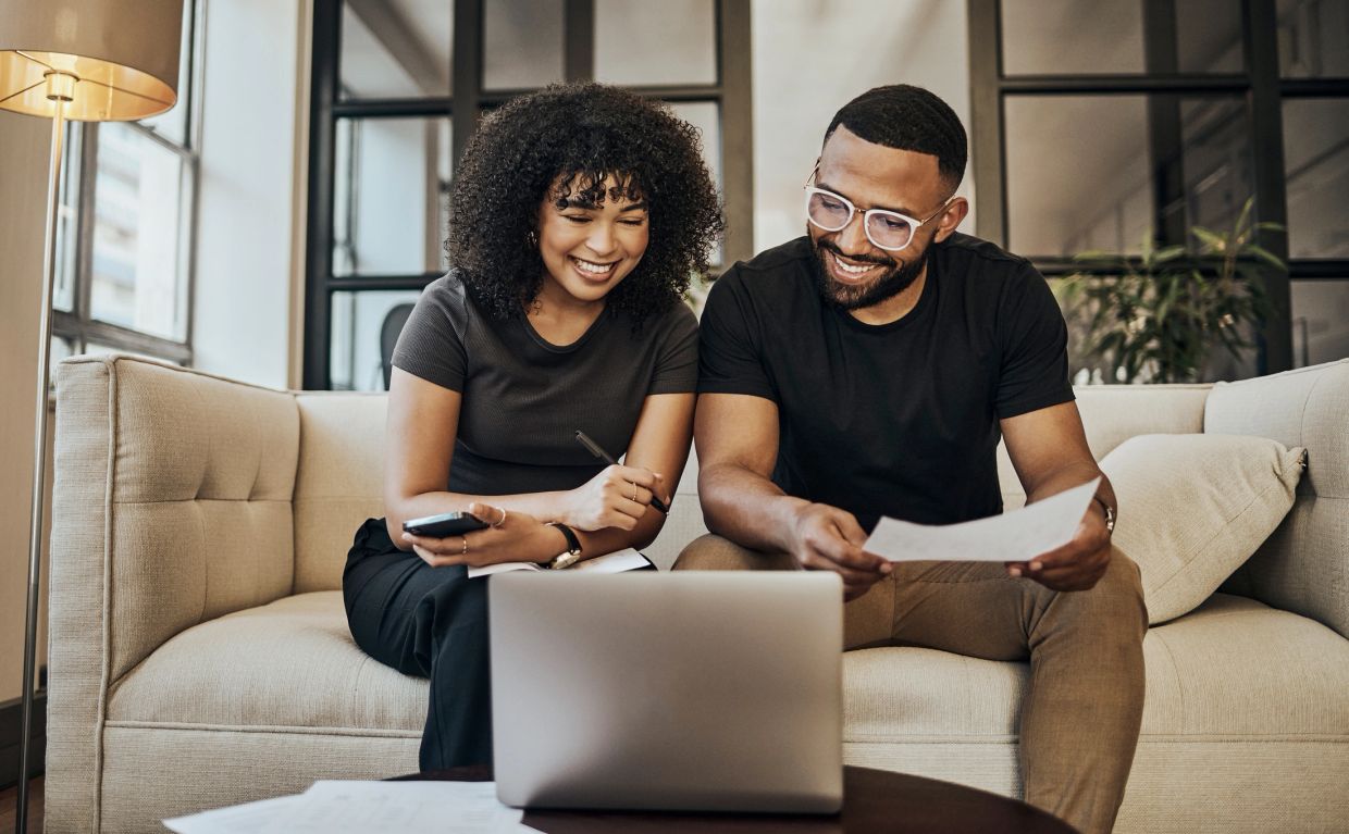 A happy couple sitting on a couch, reviewing documents and using a laptop.