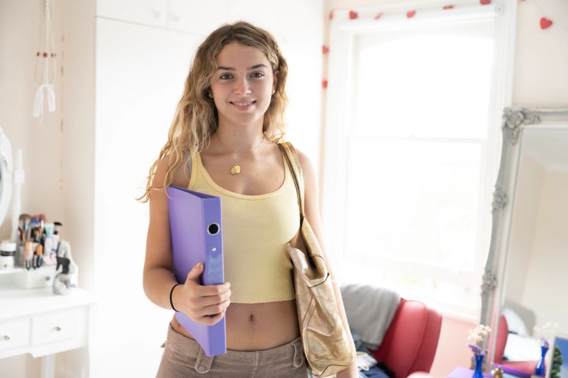 Waist-up front view of longhaired Caucasian girl standing in naturally lit room carrying shoulder bag, ring binder, and smiling at camera. Ready for school.