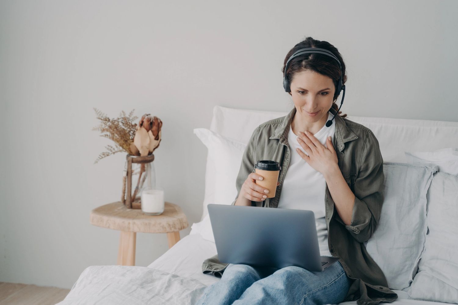 Woman with headset and coffee working on laptop in cozy room.