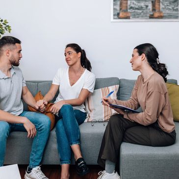 Couple holding hands during a therapy session with a counselor taking notes.