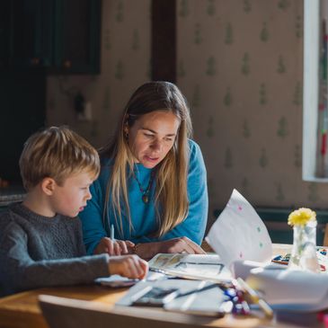 A woman helps a young boy with homework at a sunlit kitchen table.