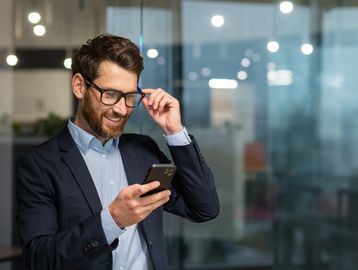 Smiling businessman adjusting glasses while looking at smartphone.