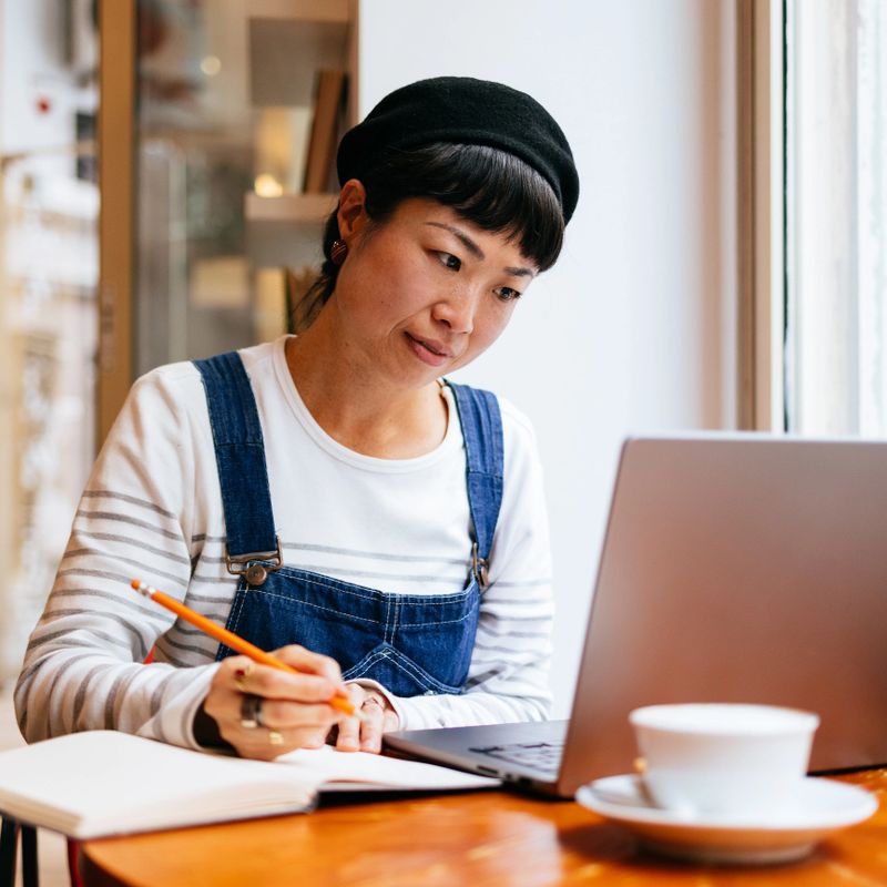 Beautiful Asian woman reading business report on her laptop and writing notes in a notebook while sitting at coffee shop desk with a cup of coffee.