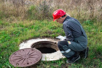 Worker inspecting an open manhole in a grassy area with a clipboard.