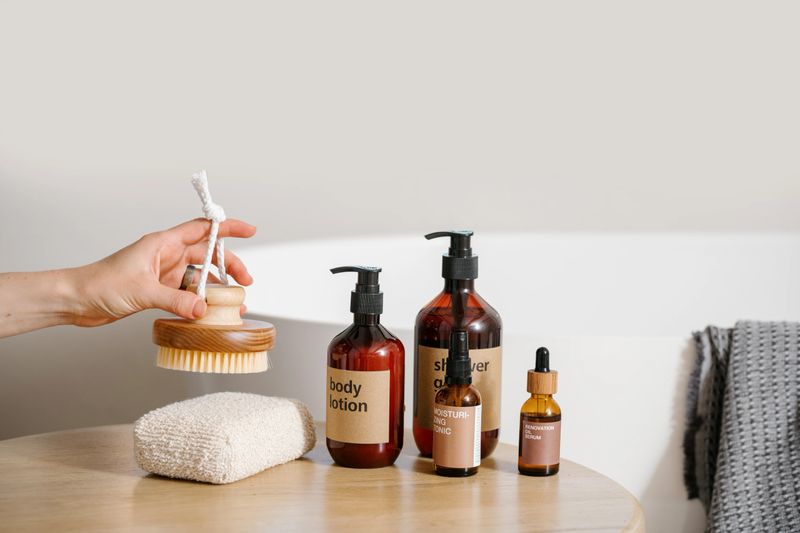 organic skincare cosmetics, brown dispenser bottles with body lotion and shower gel on table and female hand hold bamboo massage brush for peeling on white wall background