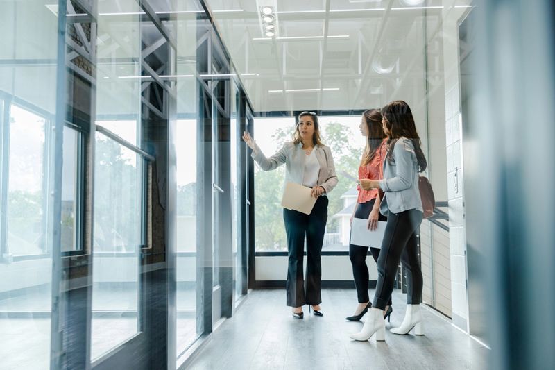 The mature adult female real estate broker gestures as she describes the office spaces to the two female entrepreneurs.