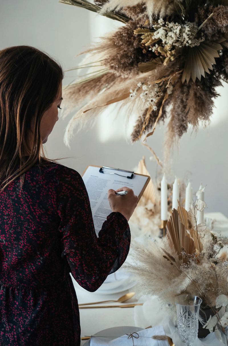 Back view of unrecognizable woman in elegant dress setting the table for new year party and writing notes on a clipboard while standing in a dinning room.