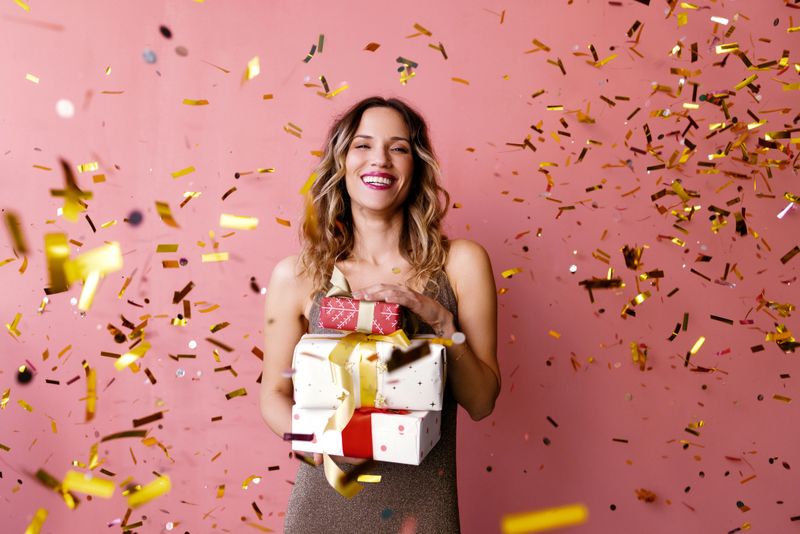 Giving is better than receiving: A portrait of a smiling Caucasian female in glitter brown dress looking excited after shopping Christmas gifts. (studio shot, pink background)