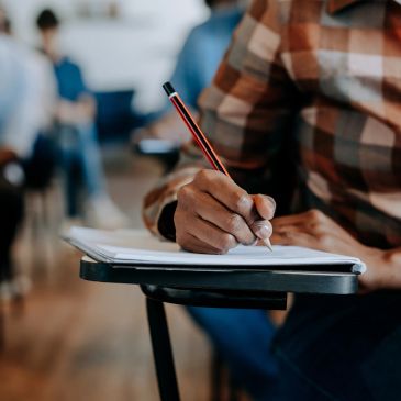 Person writing notes with a pencil in a classroom setting.