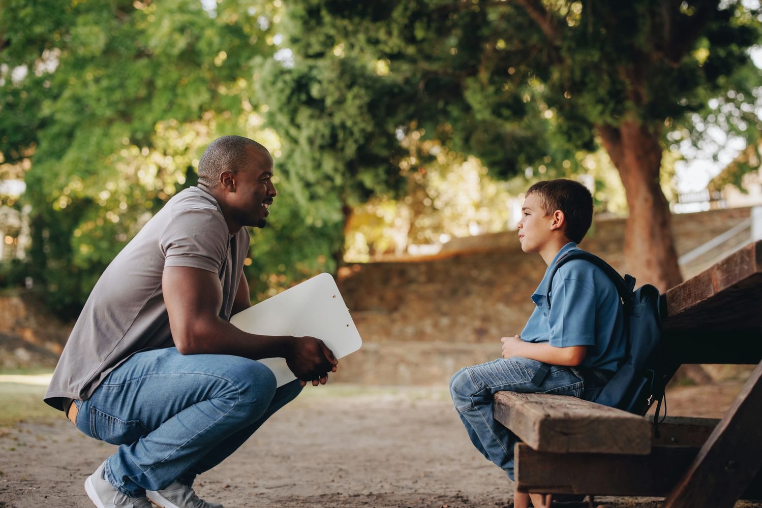 Man crouching and talking to boy sitting on a bench outdoors.