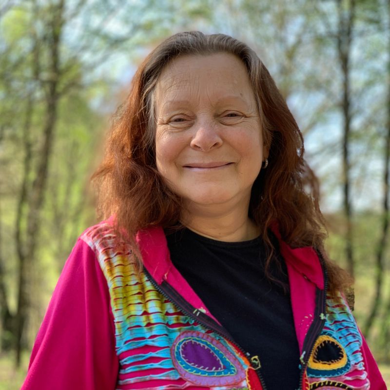 Portrait of an elderly woman. She wears a colorful vest with a lot of pink. In the background the trees of a forest blurred.