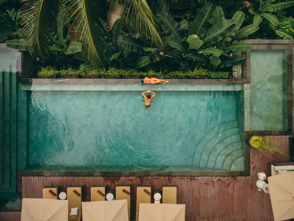A man swims and a woman sunbathes by a pool surrounded by lush greenery.