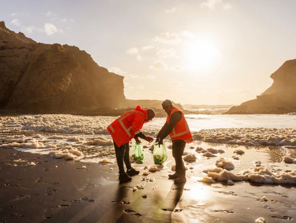 Two people in orange vests cleaning a beach at sunset.