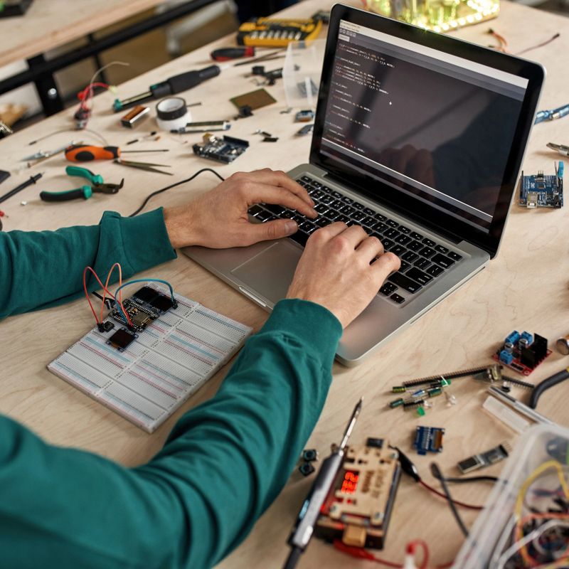 Cropped male IT technician or programmer programming code of microchip on laptop in office. Man working at table with variety tools and components. Modern technology and innovation