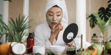 Woman in bathrobe applying green facial mask while looking in a mirror.