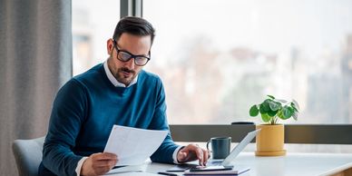 Man in glasses reviewing documents at a desk with a laptop and coffee.