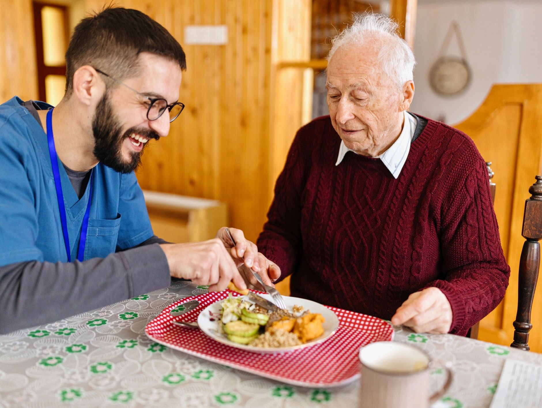 A caregiver happily assists an elderly man with his meal at a dining table.