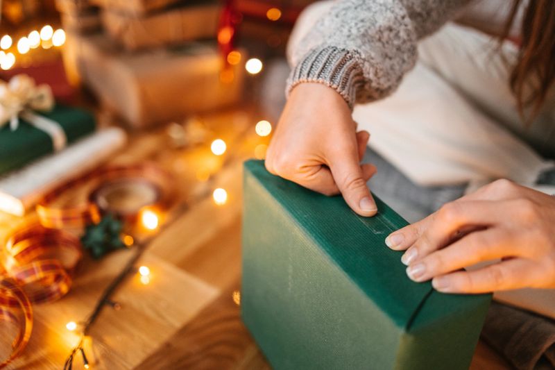 A top view of a young woman wrapping Christmas present in green wrapping paper next to a Christmas tree. Sticking paper with a tape