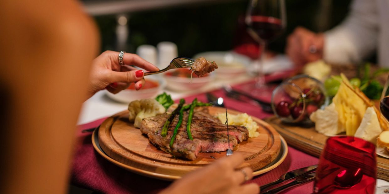 Person eating grilled steak with asparagus and mashed potatoes at a dinner table.